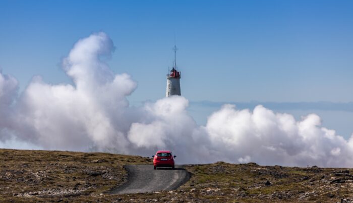 A red car is heading lighttower, that is in distance hidden partially behind white clouds